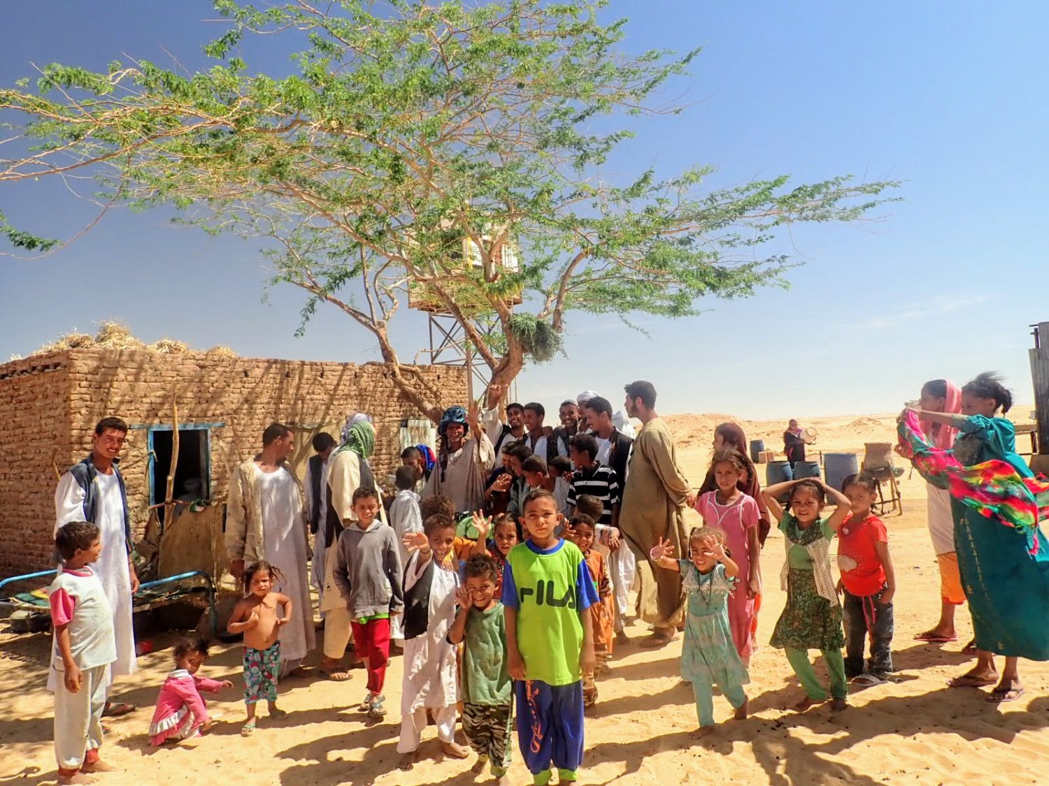 Large family outside mud hut in Sudan desert waving and smiling