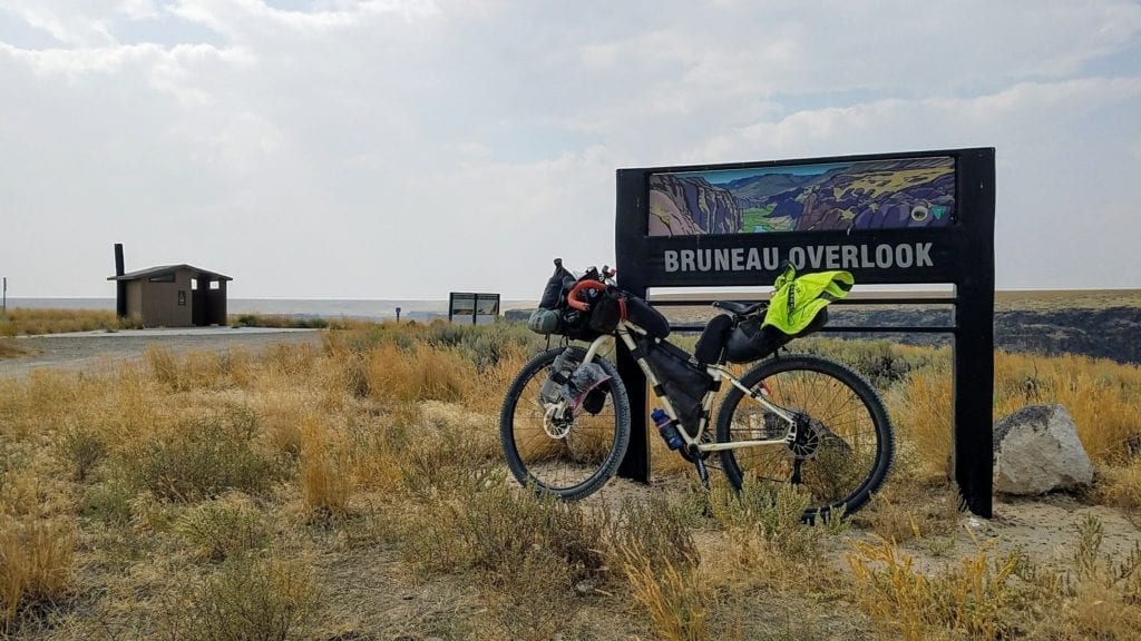 Sign says Bruneau Overlook with bicycle leaning against it, and parking area in background